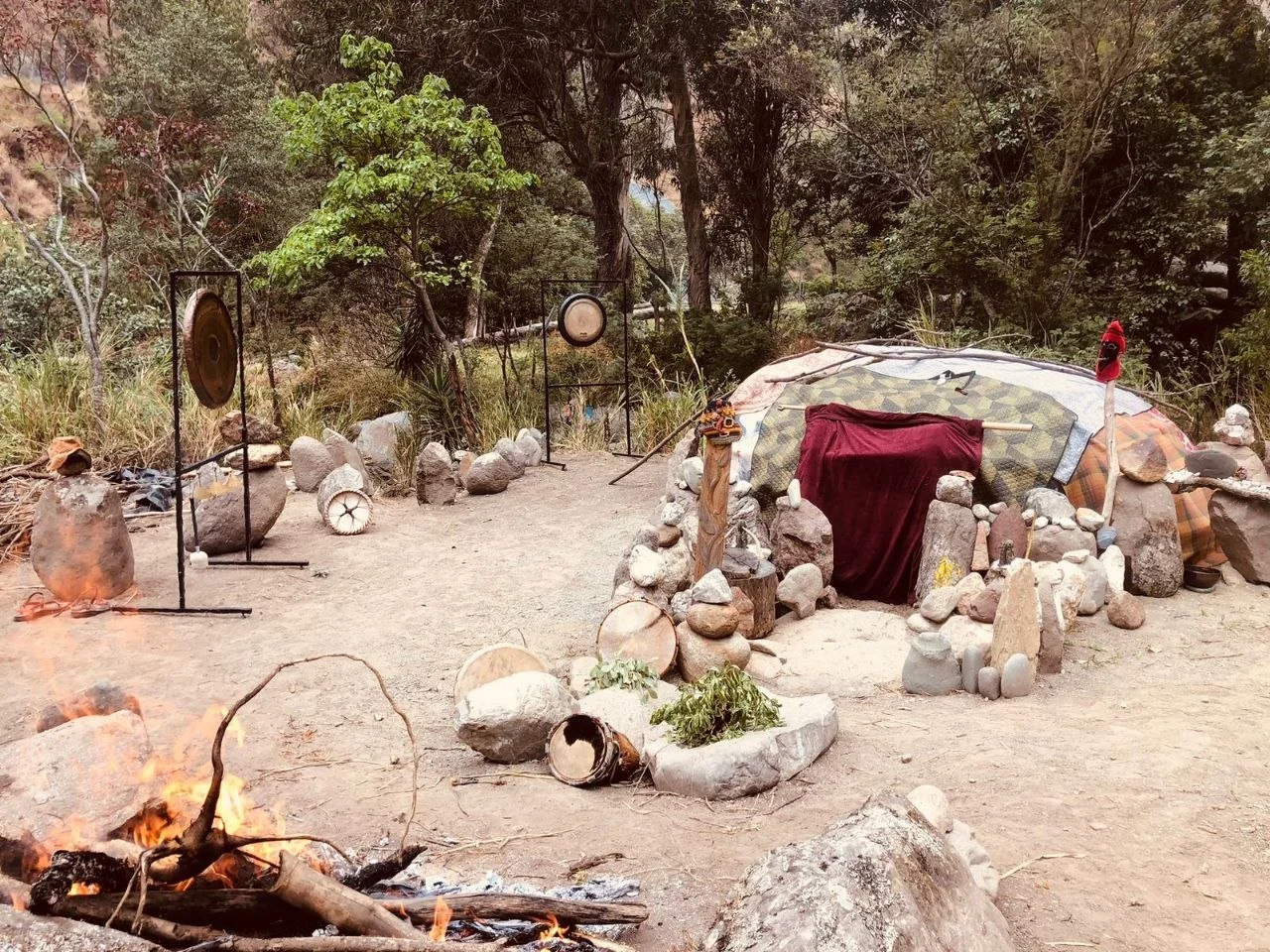 An outdoor scene with a fire in the foreground, surrounded by rocks and pottery, and a large rock shelter covered with fabric in the background. There are trees and natural scenery around.