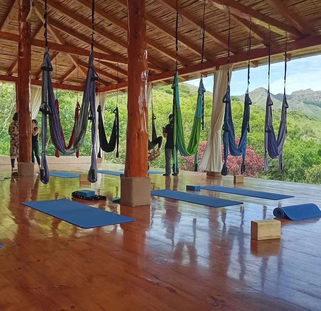 Aerial yoga studio with yoga mats, aerial hammocks, and wooden blocks, set against a backdrop of lush green mountains.