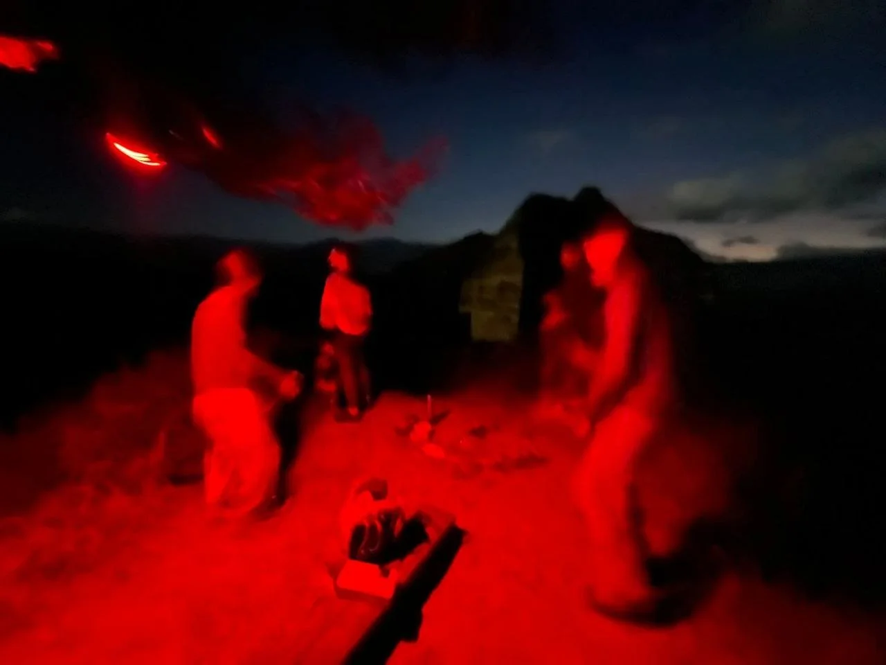 Three people gathered outdoors at night around a small fire, illuminated with a red glow. The background shows a dark sky with some clouds and distant mountains.