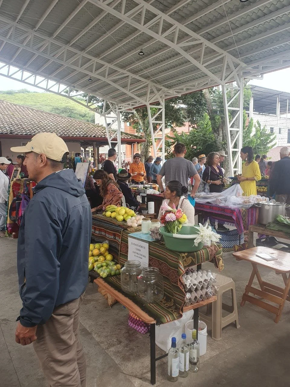 A bustling outdoor market under a covered structure with vendors selling various goods, including fresh produce, flowers, and bottled drinks, surrounded by shoppers.
