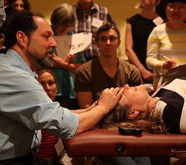 A man performing a head or face treatment on a woman lying on a treatment table, surrounded by a group of onlookers in a room.
