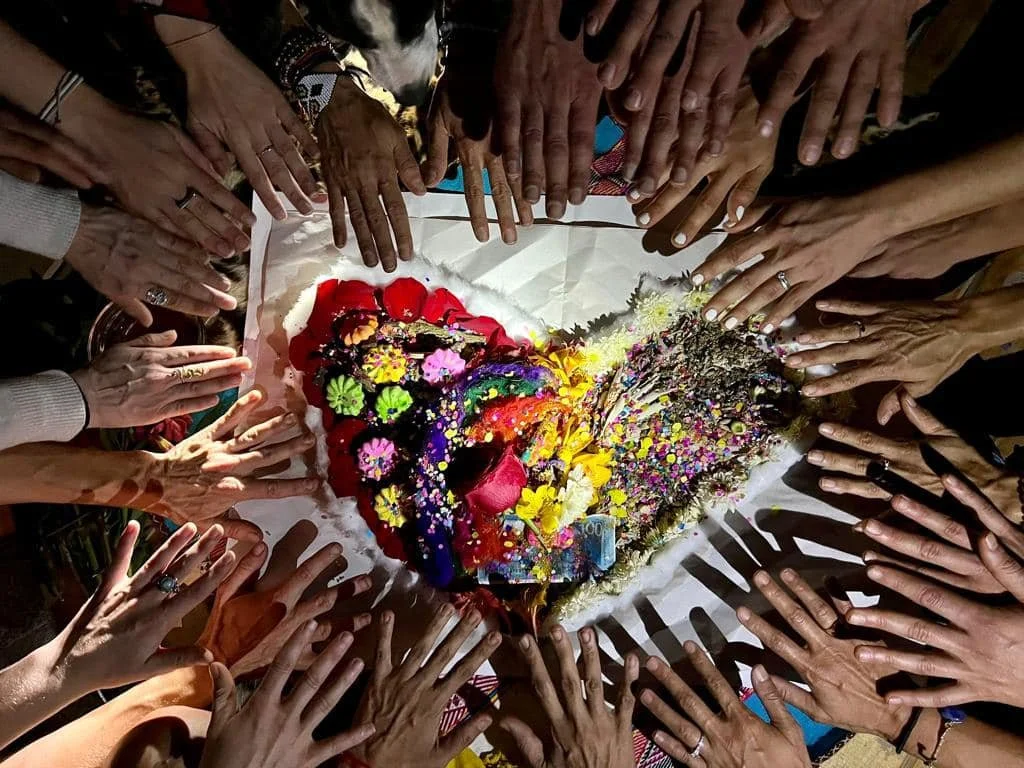 Multiple hands reaching towards a heart-shaped arrangement of flowers and various colorful items.