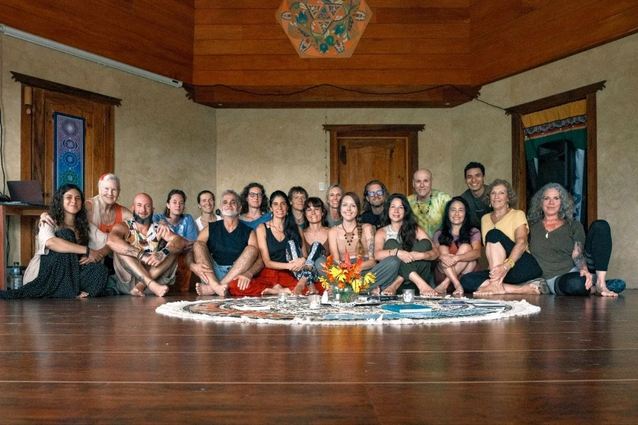 Group of people sitting on the floor in a circle inside a room with wooden walls, a central floral arrangement, and colorful curtains.