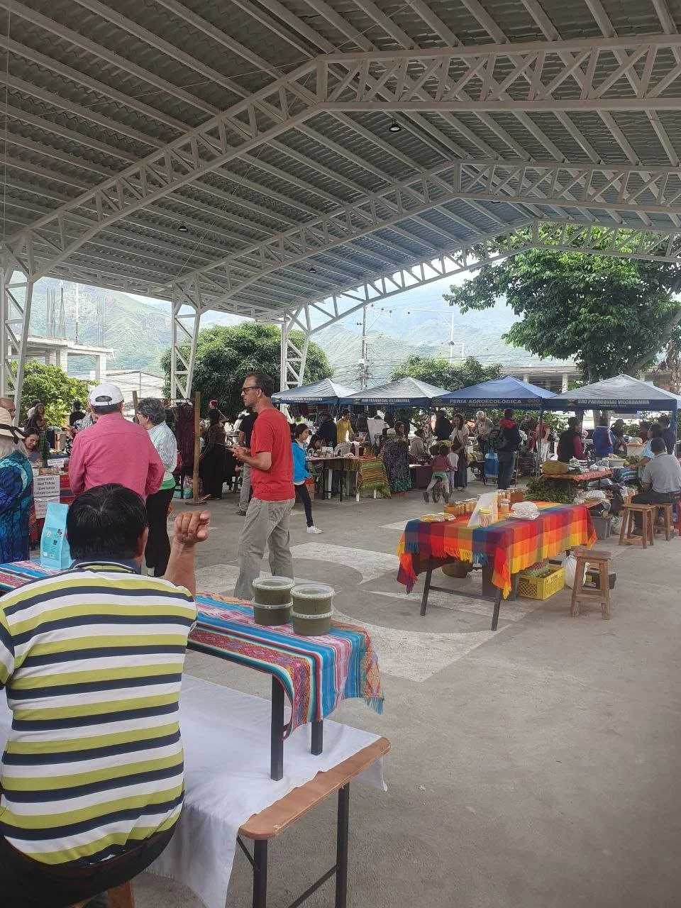 People browsing stalls at an outdoor market under a large covered structure, with vendors selling goods and tables with colorful tablecloths.