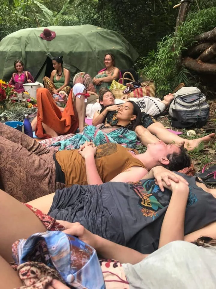 Group of people relaxing on a forest floor, lying on blankets and mats, surrounded by lush greenery and trees.