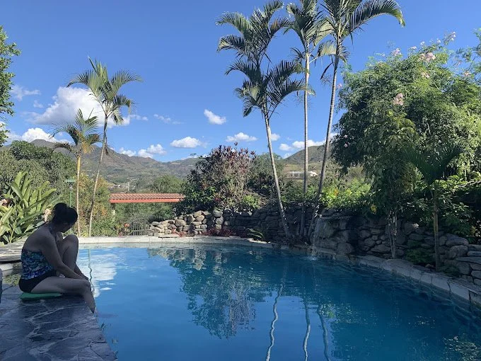 Woman sitting by a swimming pool surrounded by palm trees and lush green plants with mountains and blue sky in the background.