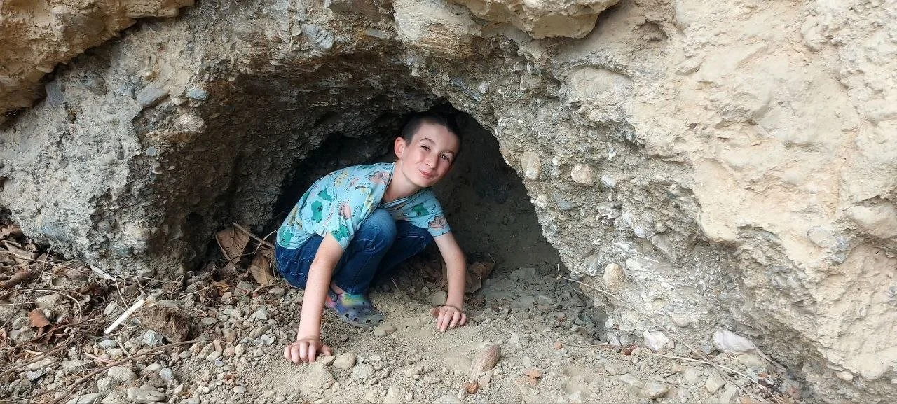 A young boy crawling through a small rocky tunnel in a dirt area outdoors.