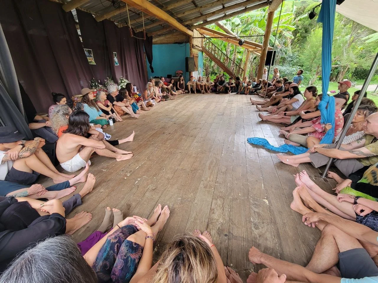 People sitting in a large circle on a wooden floor inside a rustic open-air pavilion with forest in the background, participating in a group gathering or event.