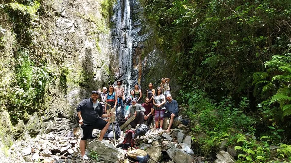 Group of people posing in front of a waterfall surrounded by lush green foliage on rocky terrain.