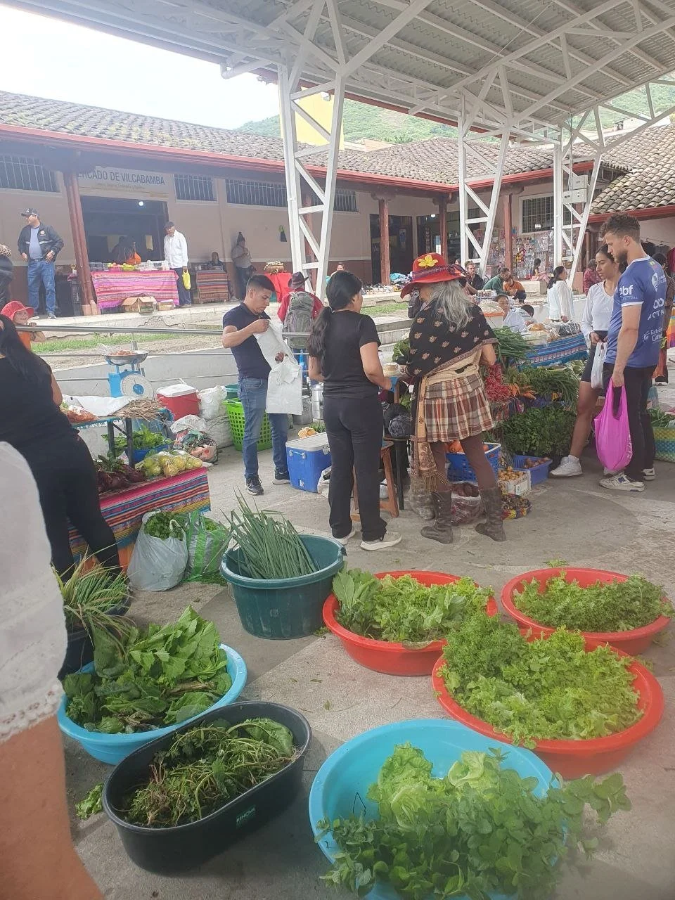 Market scene with fresh vegetables in large bowls and people shopping under a roofed area.
