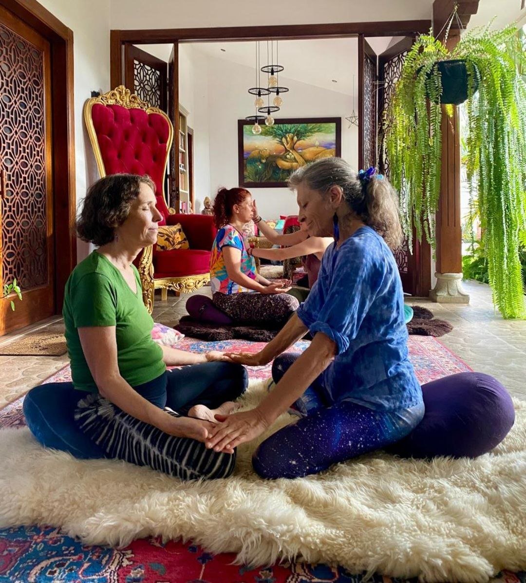 Women participating in a meditation or spiritual workshop, sitting on a colorful rug with their legs crossed in a cozy, decorated room with plants, artwork, and a large red throne-like chair in the background.