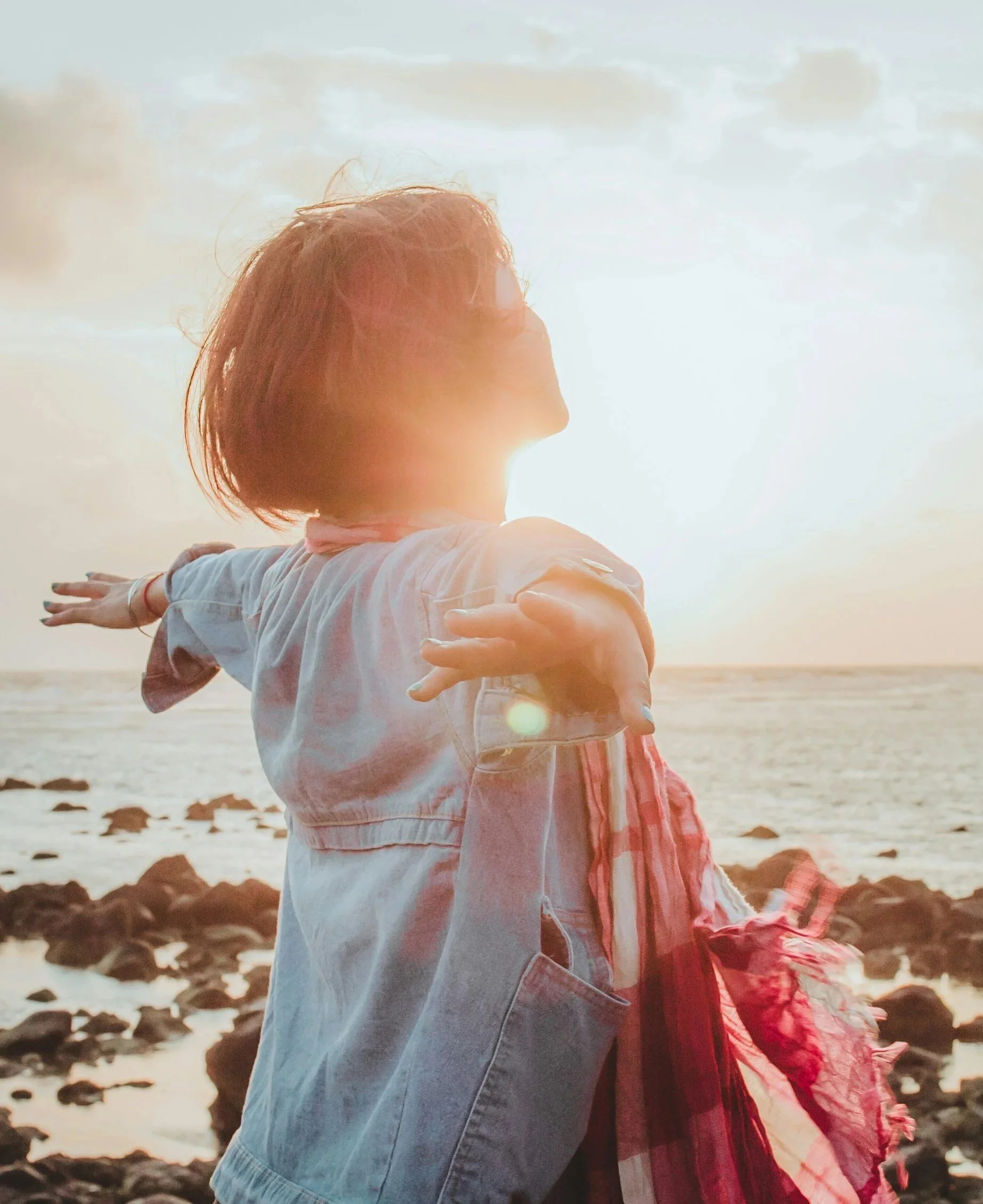 Two children are hugging on a rocky beach during sunset or sunrise, with one child wearing a denim jacket and the other in a red jacket and pink scarf. The sky is partly cloudy, and the sunlight creates a warm glow.