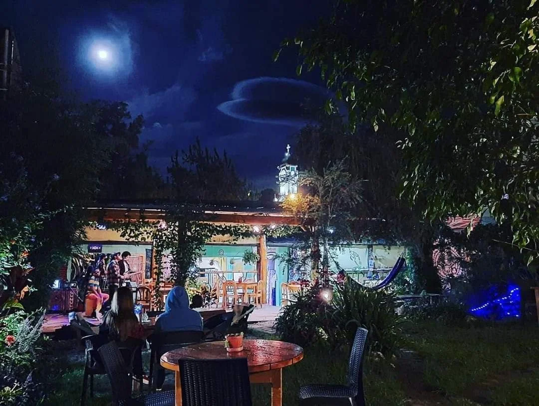 People sitting around a lit table outside at night, with a small stage and a house with a clock tower in the background.