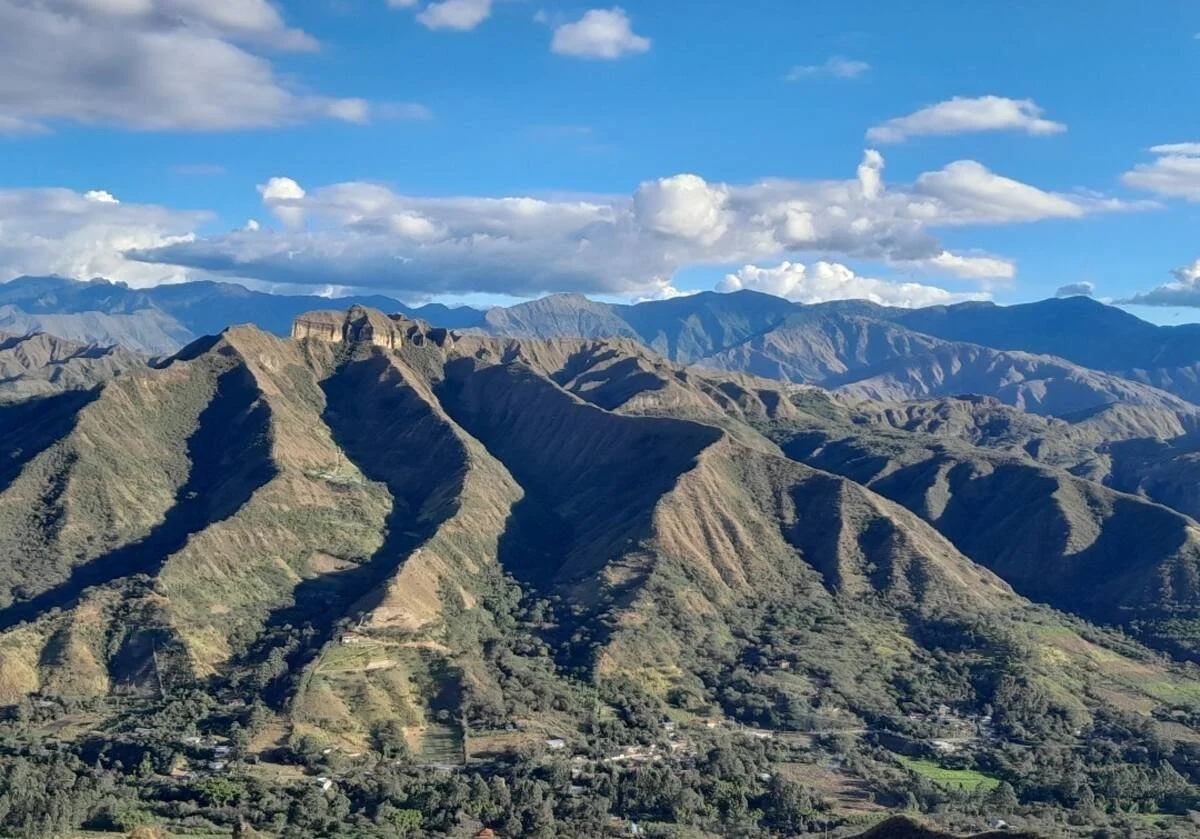 Scenic view of green, rugged mountains with terraces and scattered trees against a partly cloudy blue sky.