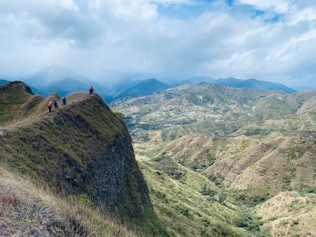 Group of hikers walking along a ridge in a mountainous landscape with green hills and cloudy sky in the background.