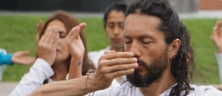 A group of people participating in a spiritual or meditation practice outdoors, with one man in the foreground with long hair and beard, eyes closed, as someone touches his face gently.