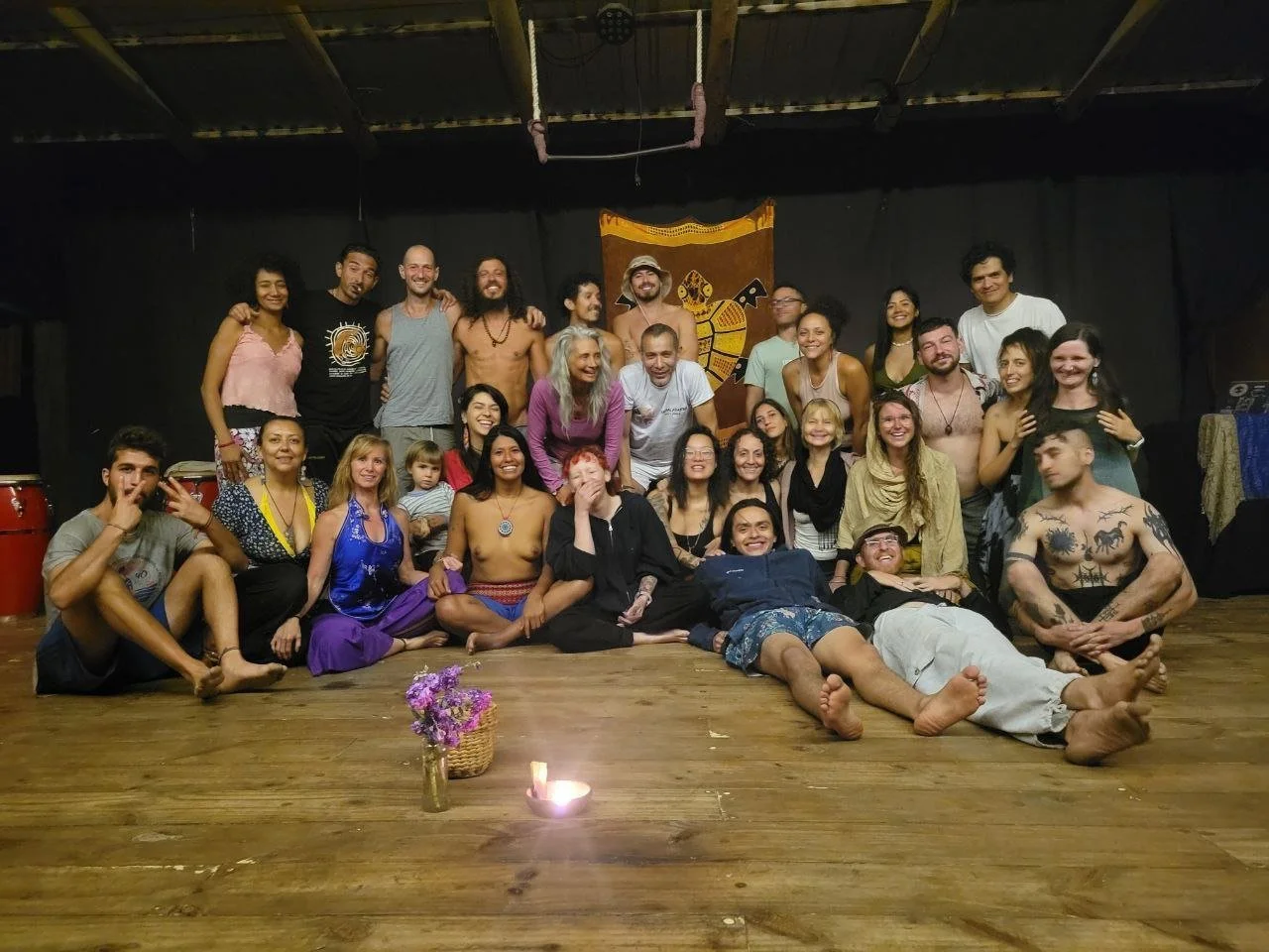 Group of people gathered on a wooden floor for a group photo, some sitting, some kneeling, and some standing, with a black backdrop and a decorative tapestry featuring a stylized animal. A small bouquet of purple flowers and a lit candle are in the f