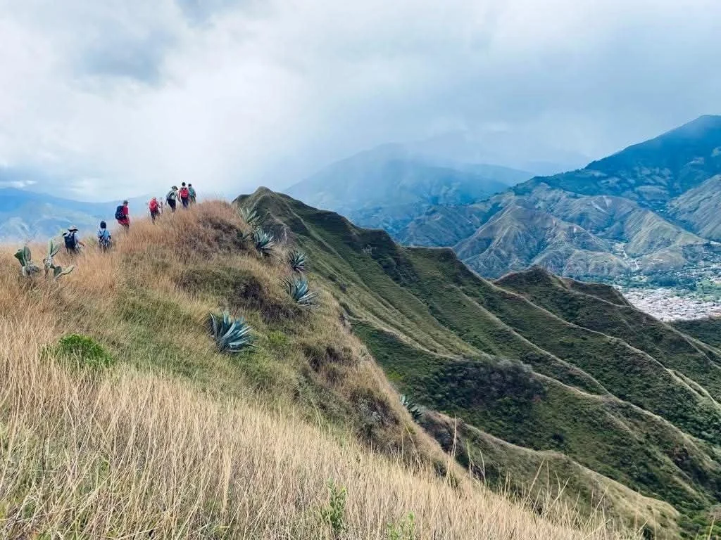 Hikers walking along a mountain ridge with grassy slopes and agave plants, overlooking a valley and distant mountains under a cloudy sky.