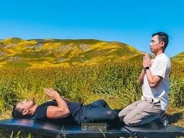 Two people practicing yoga outdoors in a field under a blue sky, one lying down and the other kneeling.