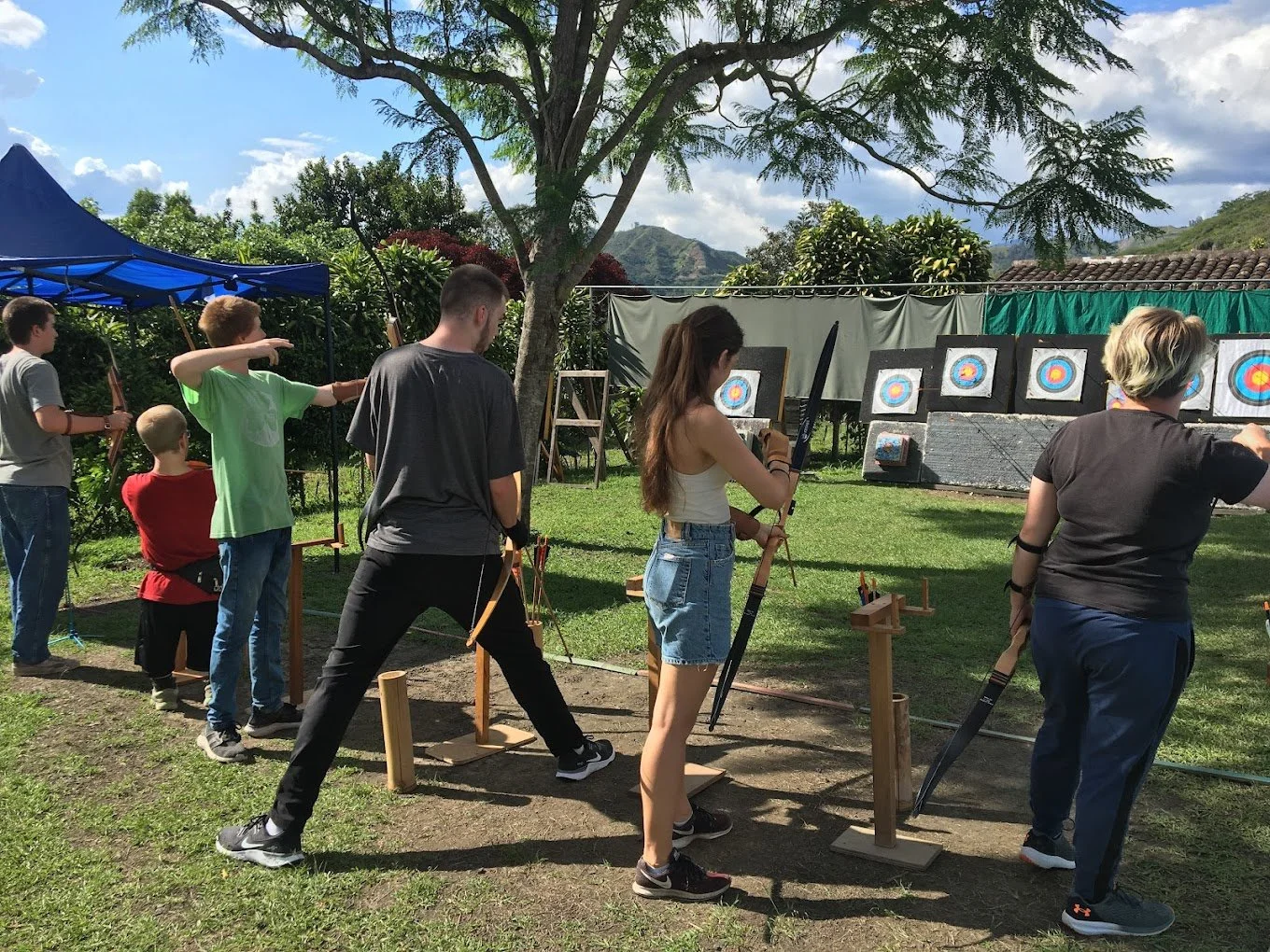 People participating in an outdoor archery activity, aiming at targets, under a large tree with a fence and landscape in the background.