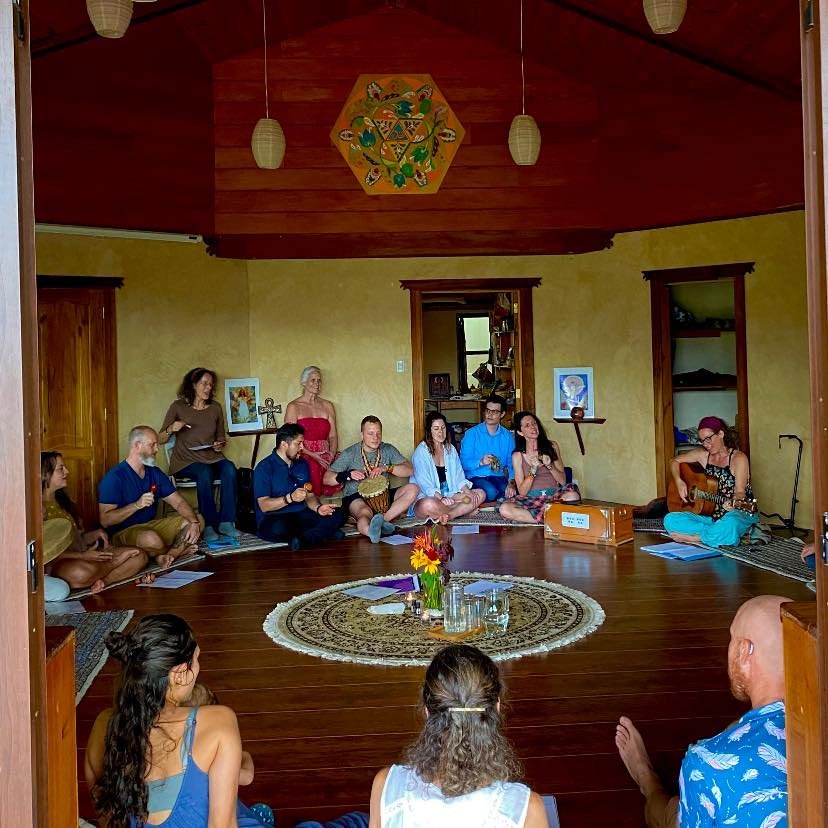 Group of people sitting in a circle on the floor, with some playing musical instruments and others singing, in a cozy room with wooden walls and a decorative ceiling, during a gathering or musical session.