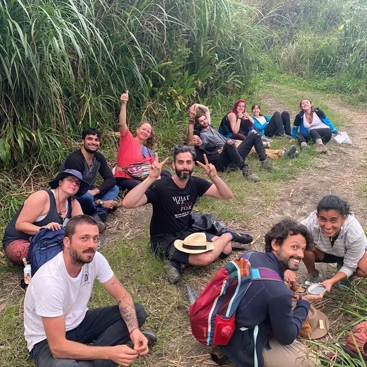 Group of ten friends relaxing and posing on a grassy trail next to tall green plants in a natural outdoor setting.