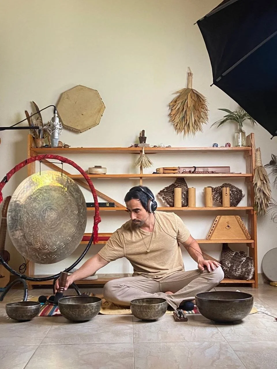 A man sitting cross-legged on the floor with headphones, playing Tibetan singing bowls during a recording session in a room with musical instruments and decorative objects on a wooden shelf.