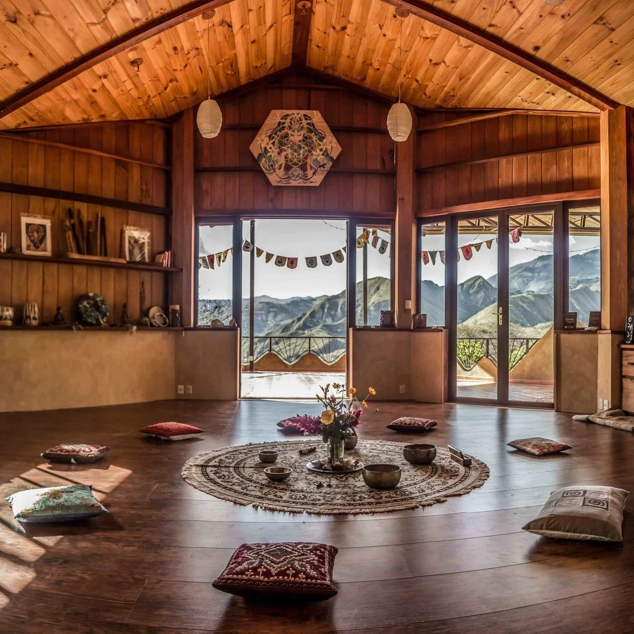 Indoor wooden room with a view of mountains, decorated with cushions, a circular rug, and a centerpiece of flowers, with sliding glass doors leading to a balcony.