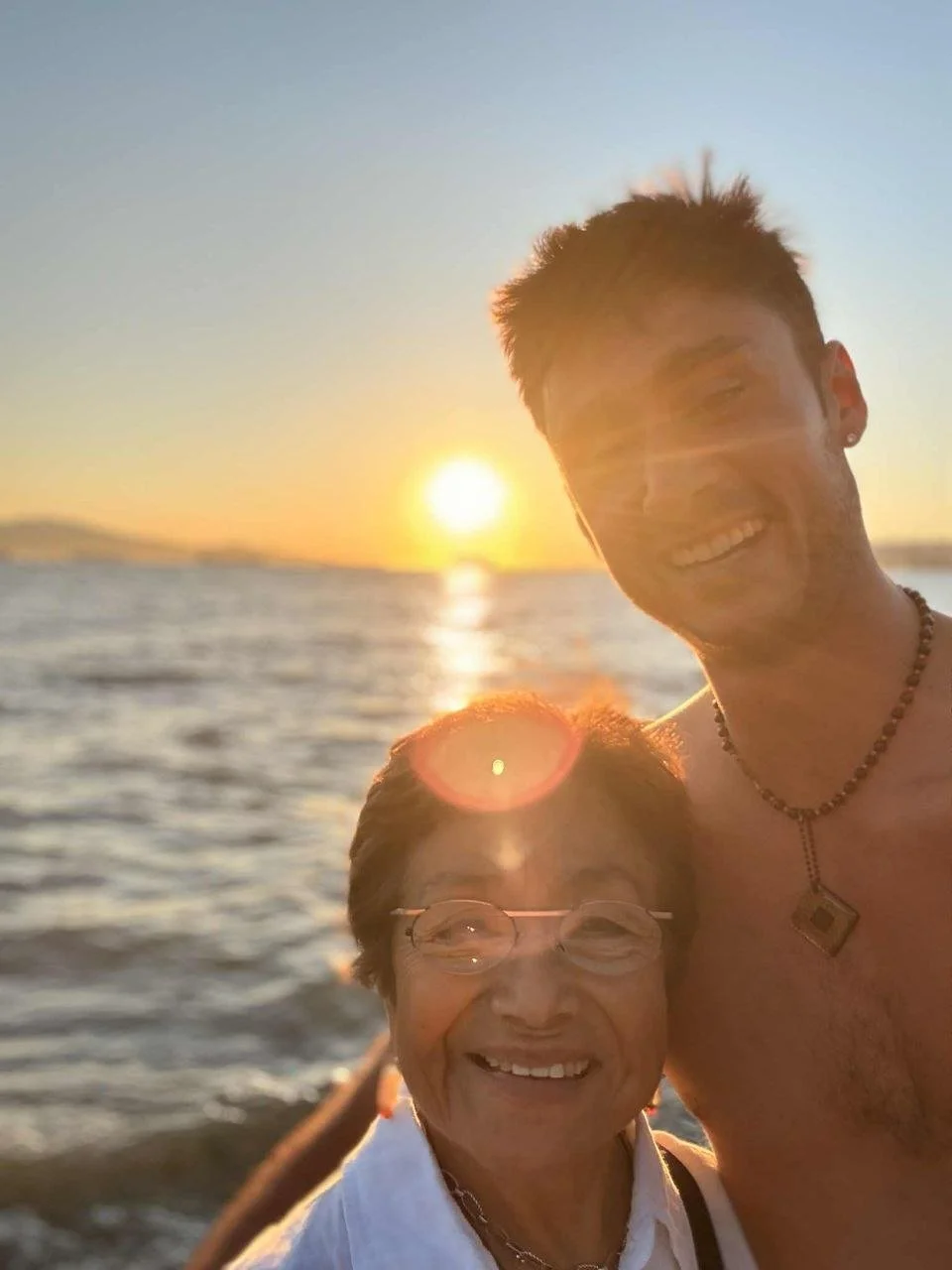 A young man and an older woman taking a selfie together on the beach at sunset, both smiling.