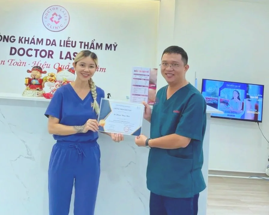 A woman in blue medical scrubs receiving a certificate from a man in green medical scrubs at a skin care clinic, with decorations and a TV displaying a presentation in the background.