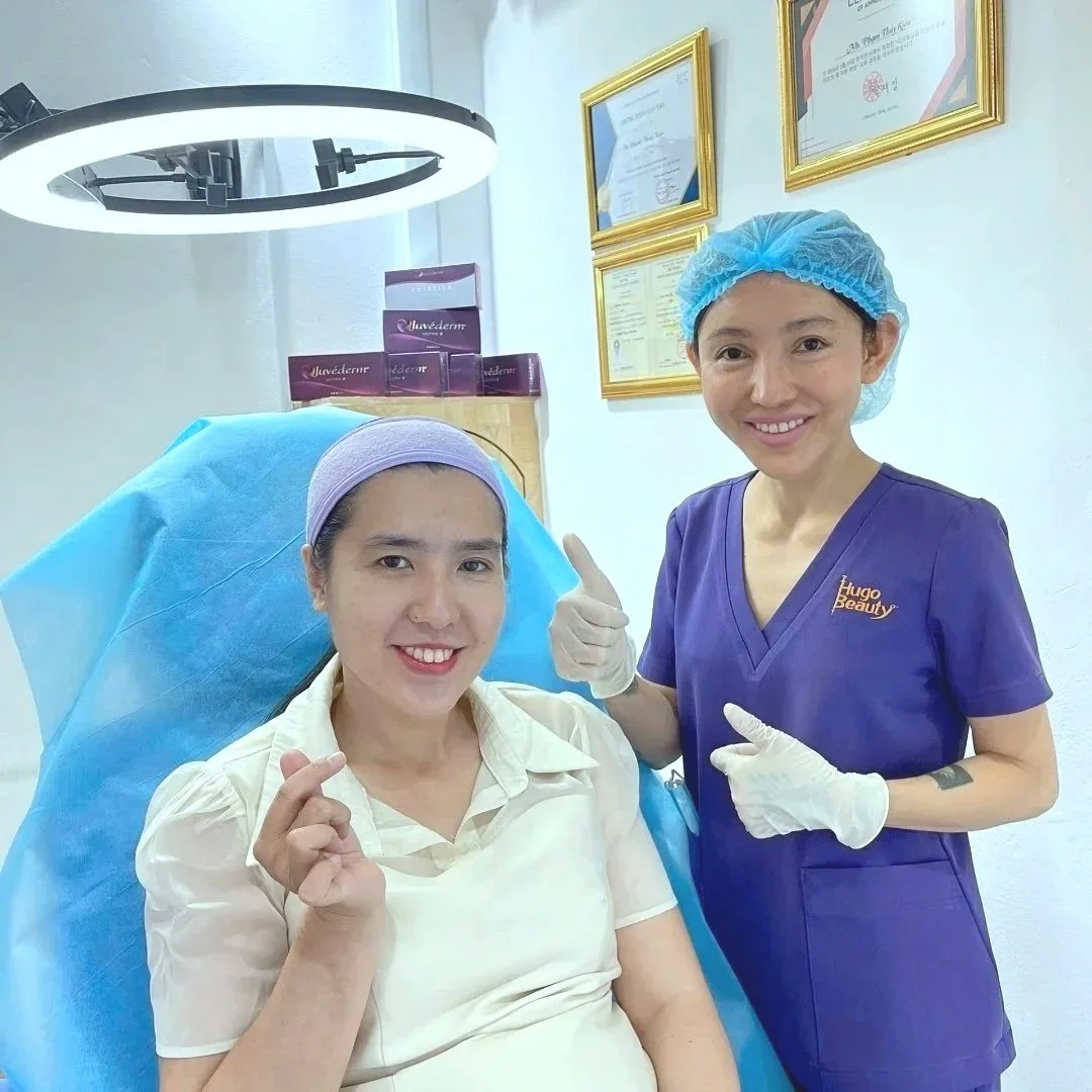 A woman in a white shirt seated in a medical chair with a blue cover, smiling while making a heart gesture with her right hand. Next to her, a nurse or cosmetologist in a purple uniform, with a blue hair cover and white gloves, is standing and smiling, pointing at the woman. Behind them, framed certificates and boxes of skincare products are visible, with a large ring light overhead.