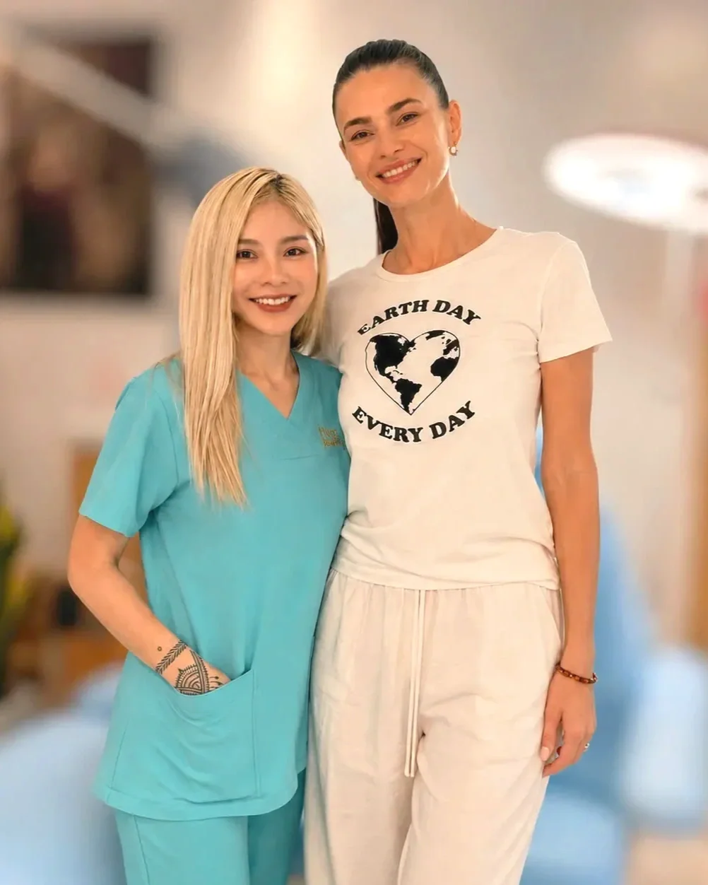 Two women smiling, one in medical scrubs and the other in casual wear with an Earth Day shirt, standing close together indoors.