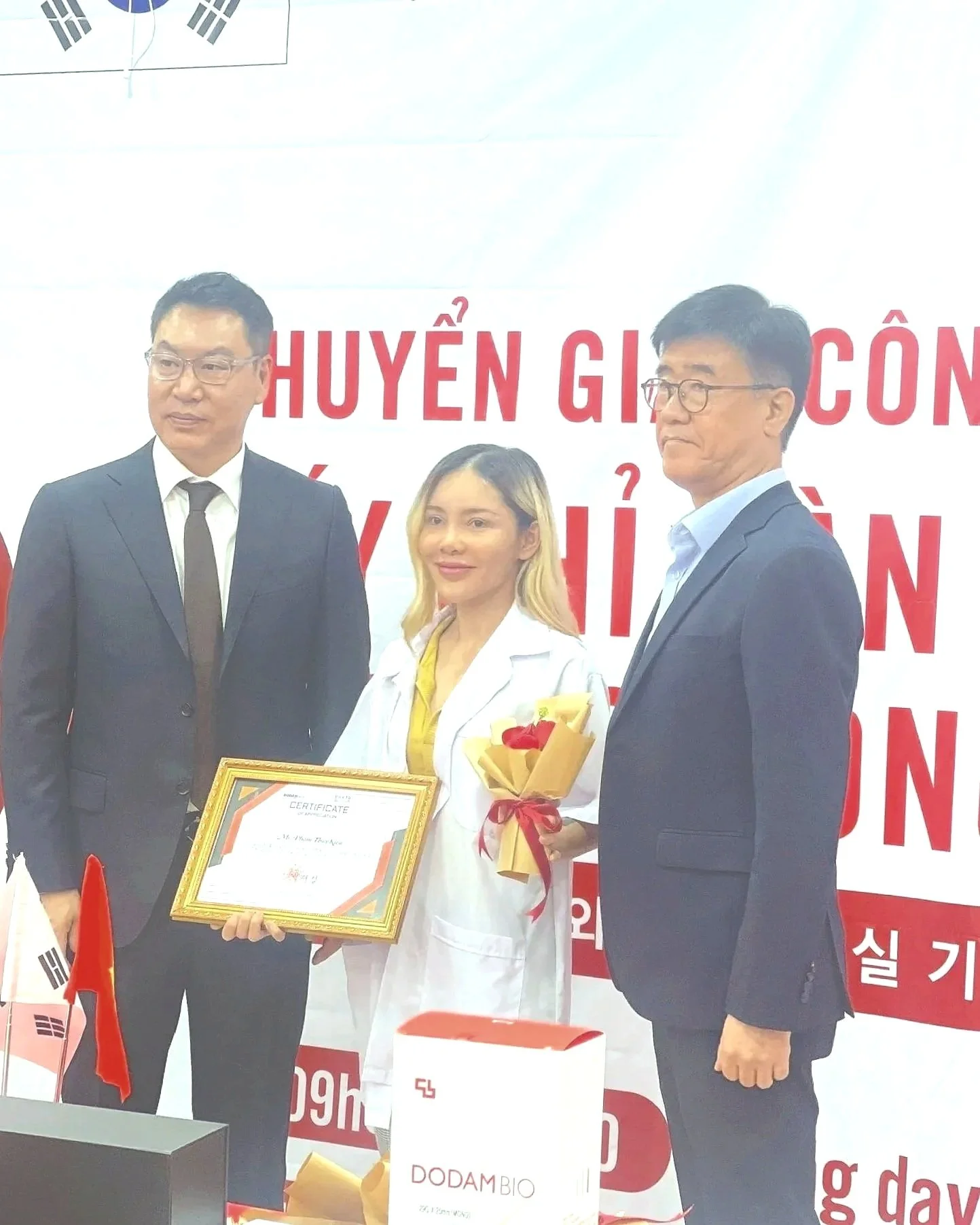 Three people standing together at an event, with two men in suits and a woman in a white coat holding a framed certificate and a bouquet of flowers. Behind them is a backdrop with red text. Flags of South Korea and Vietnam are visible on the table in front.