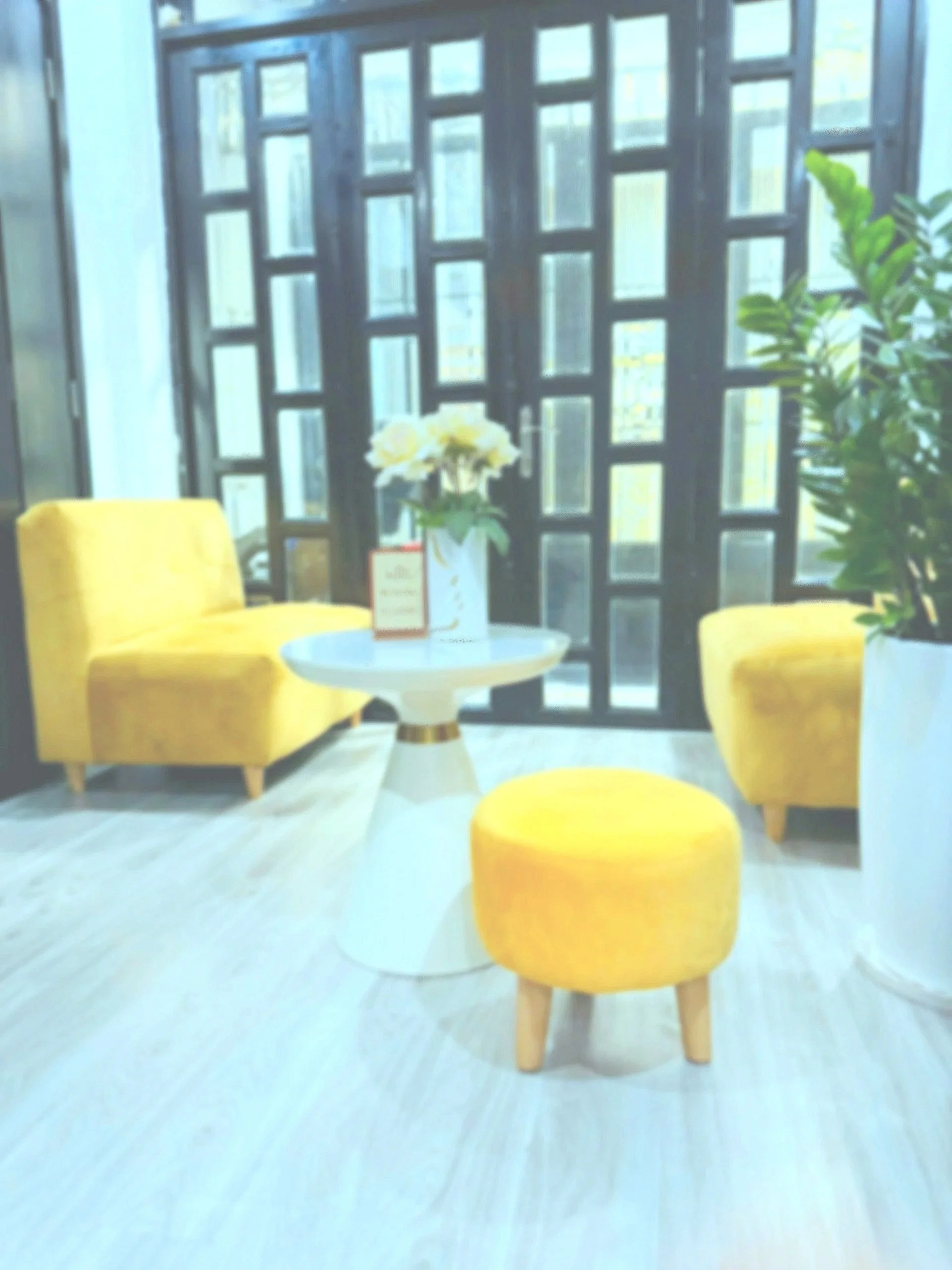 Interior space featuring two yellow upholstered chairs, a small round white table with a yellow base, a vase of white flowers, a potted green plant, and a decorative black wooden screen with geometric patterns in front of a window.
