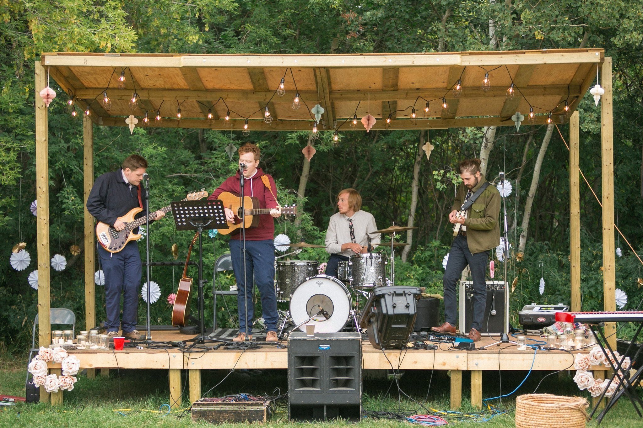 Outdoor stage with string lights, musicians performing under a rustic wooden canopy. The scene has a cozy, bohemian vibe with greenery in the background, creating an intimate setting.
