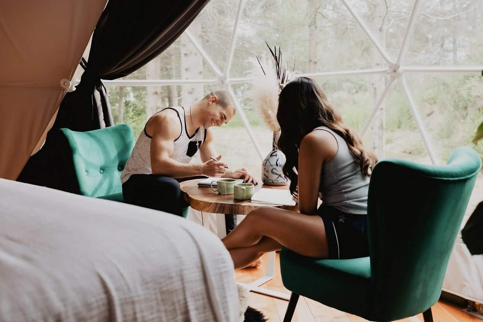 Intimate geodome setting with a couple enjoying a heartfelt conversation over a table set with delicate decor, surrounded by lush greenery outside the geometric window.