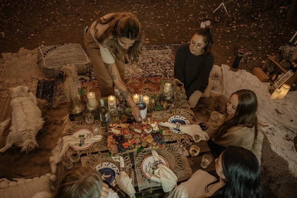 Top view of a bohemian picnic setup with friends gathered around a low table adorned with candles, charcuterie, and eclectic tableware, set in a serene forest clearing.