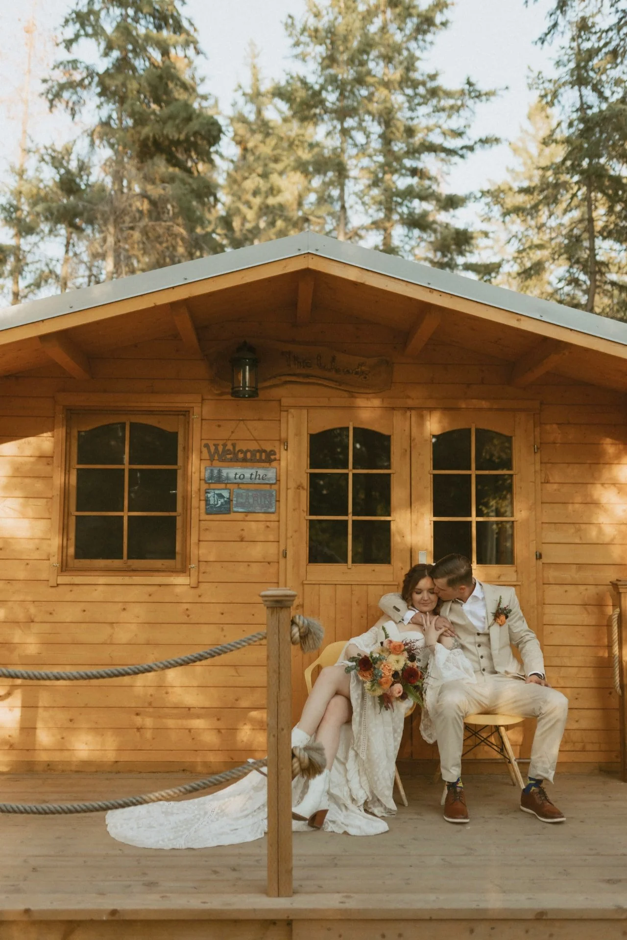 A newlywed couple relaxes on the porch of a rustic cabin labeled "Welcome to the Cabin," with the bride holding a bouquet and the groom leaning in for a tender moment.