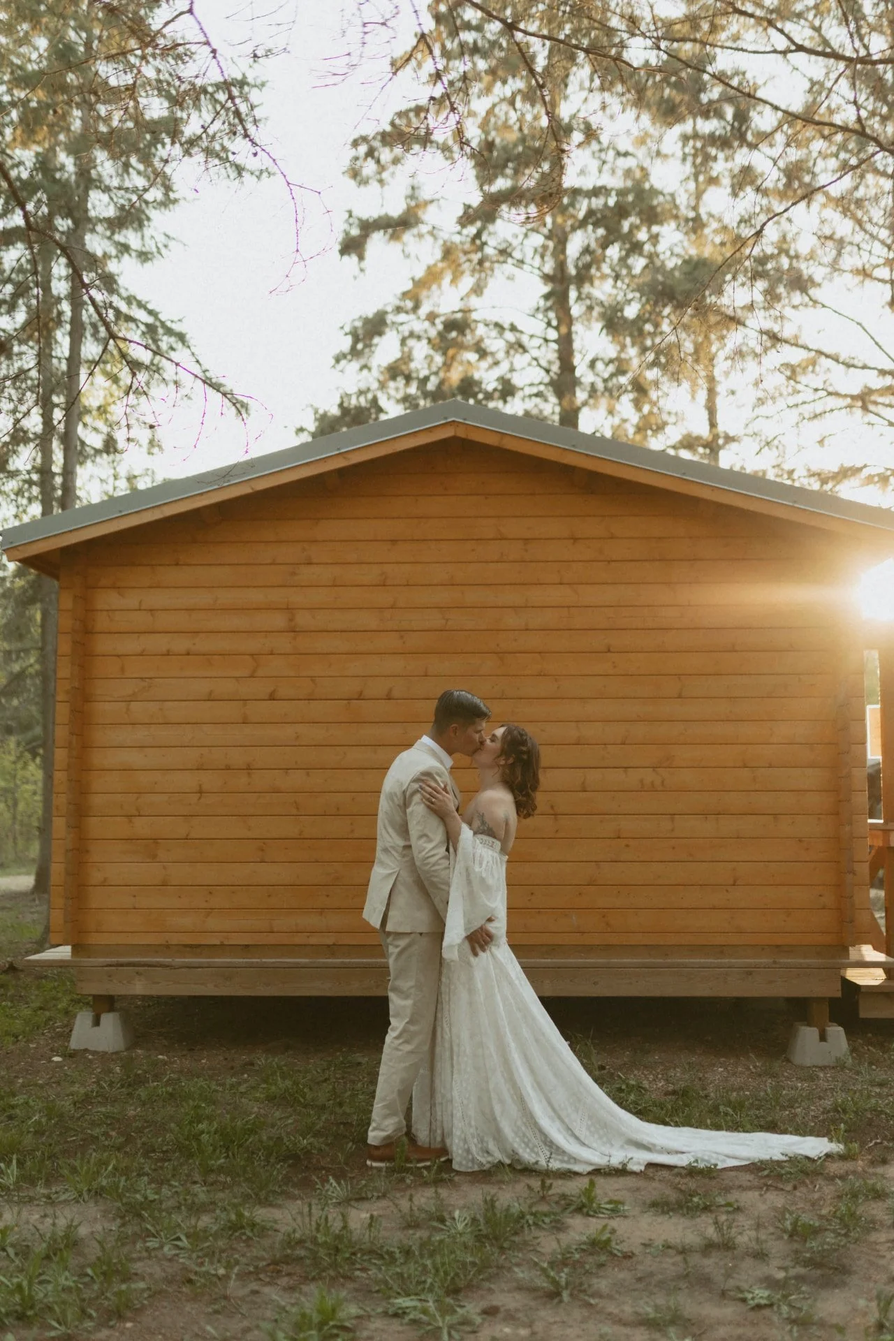 A newlywed couple shares a tender kiss in front of a rustic wooden cabin at sunset. The bride’s lace dress and the groom’s suit illuminate the serene forest backdrop, creating a romantic scene.