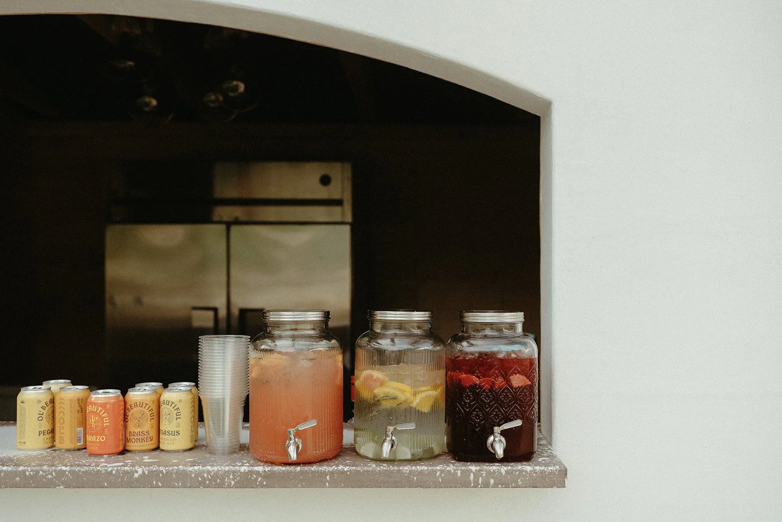 A close-up of three glass drink dispensers filled with refreshing fruit-infused beverages and an assortment of canned drinks, ready to serve guests at an outdoor retreat.