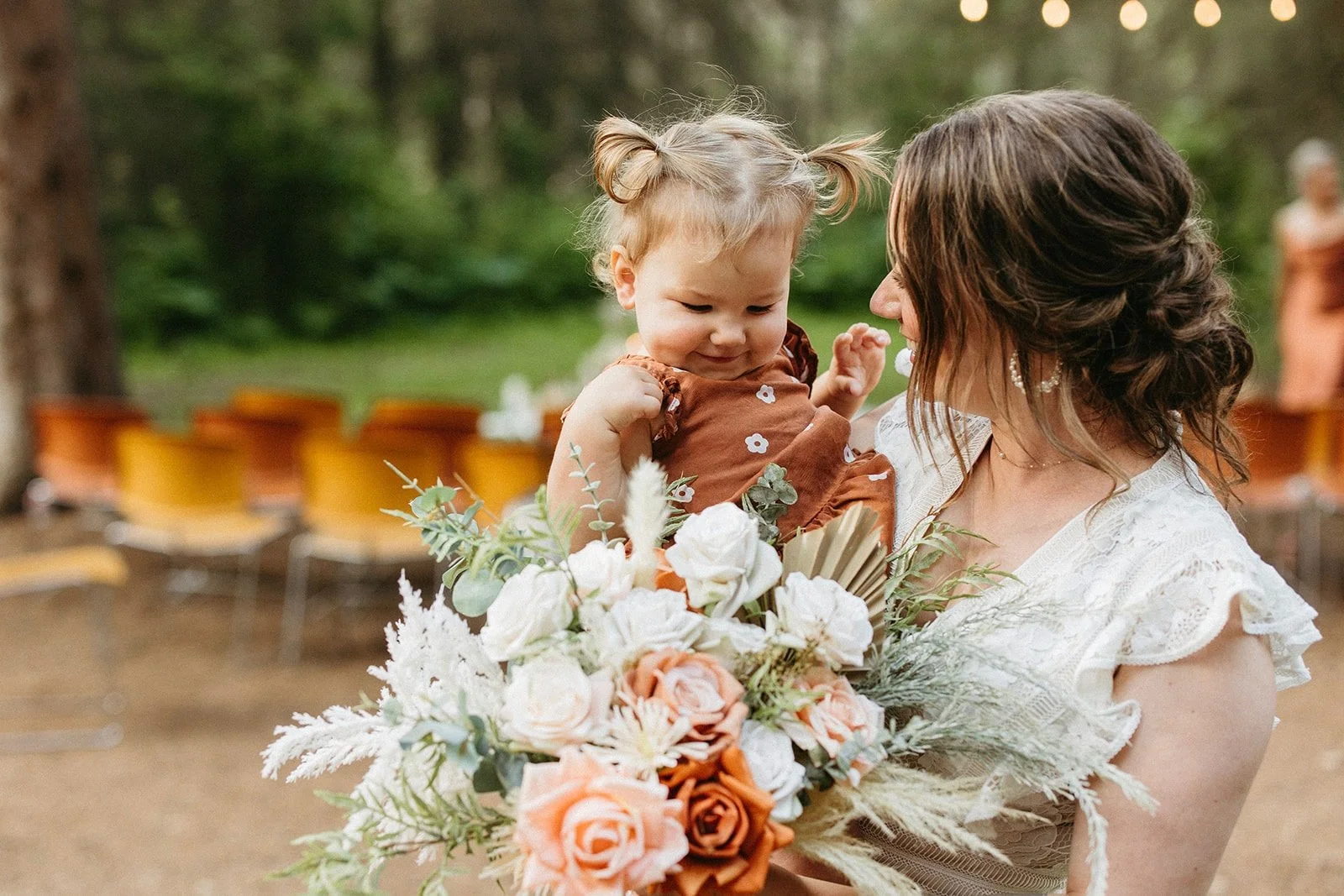 Bride holding a toddler and a beautiful bouquet, sharing a tender moment during the wedding, evoking the charm and intimacy of a boho celebration with family and loved ones.