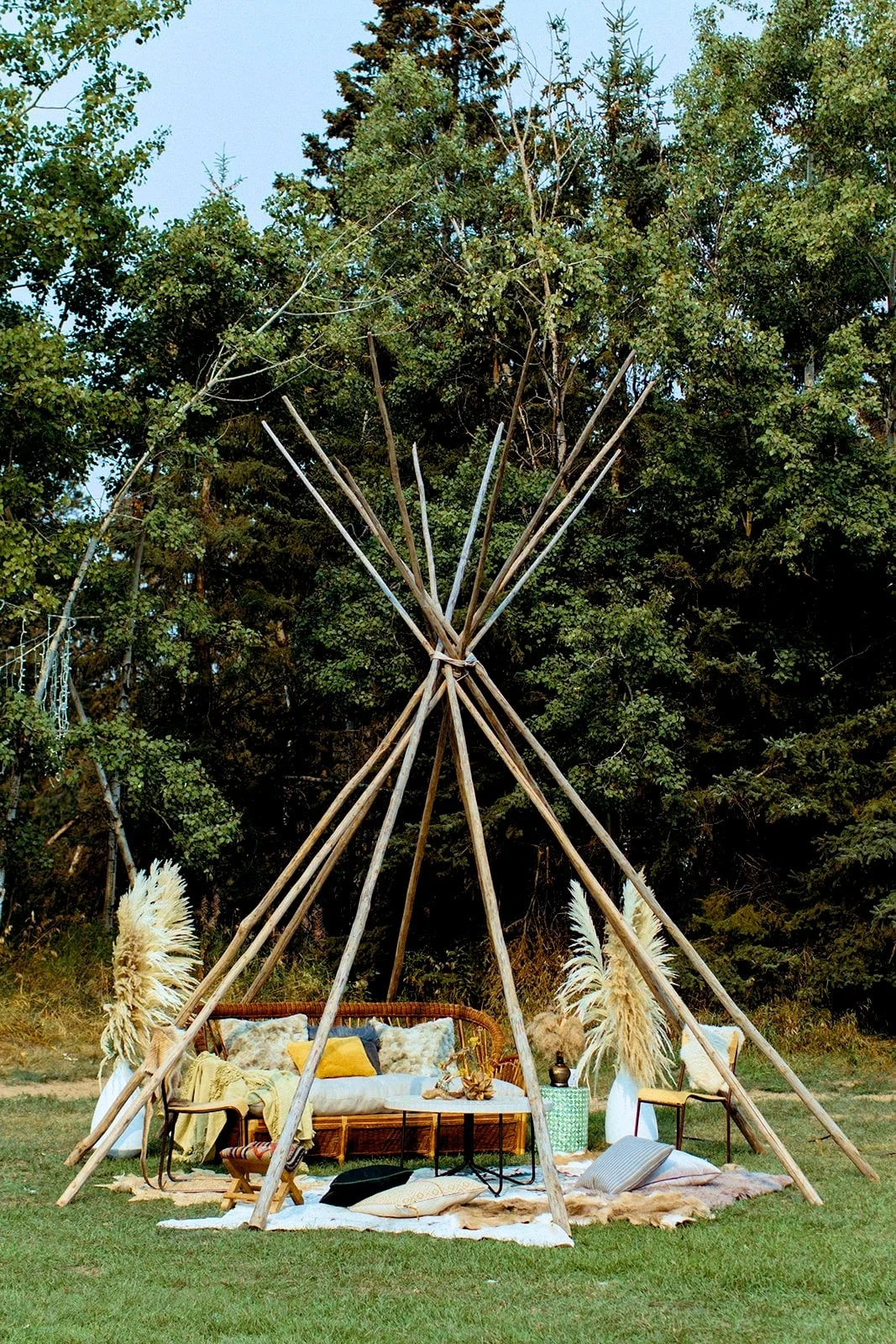 A tipi structure styled with boho-chic decor including a wicker sofa, cozy cushions, vibrant pillows, a low table, and pampas grass, nestled in a lush, green field amidst towering trees.