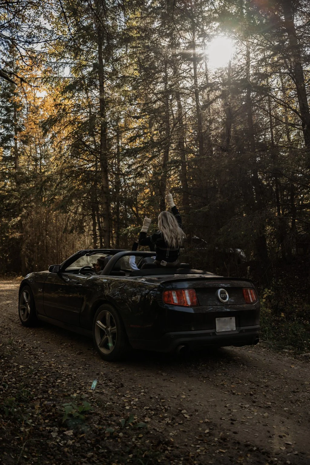 A convertible Mustang with top down on a forest trail, as a woman stands, arms raised in joy, capturing the essence of adventure and freedom amidst towering trees.