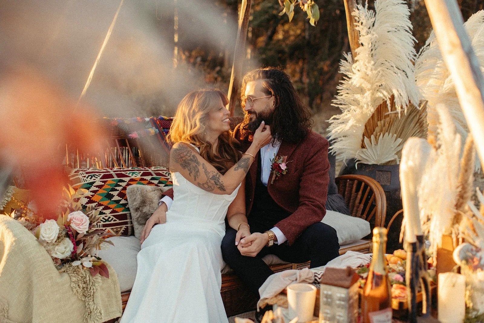 A bride and groom sit cozily in a boho-styled tipi, adorned with pampas grass and vibrant textiles, sharing an intimate moment as the sun sets, enveloping them in warm light.