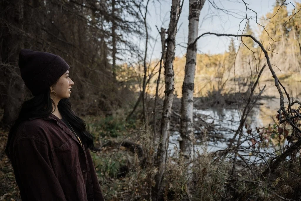A woman in a burgundy beanie and jacket stands by a serene lake, surrounded by autumnal trees. Her relaxed smile captures the essence of nature's calm.