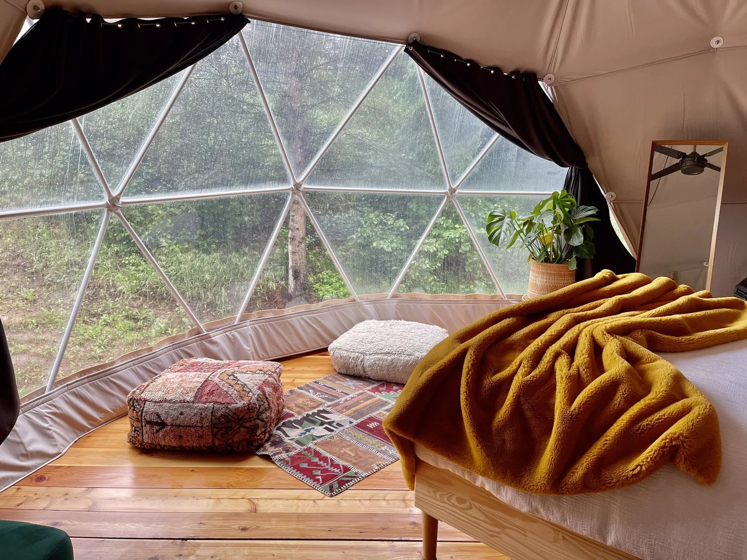 Spruce Hollow geodome interior with vibrant poufs, a lush plant, and a cozy bed draped in a golden blanket, offering tranquil forest views through a panoramic window, perfect for a stylish retreat.