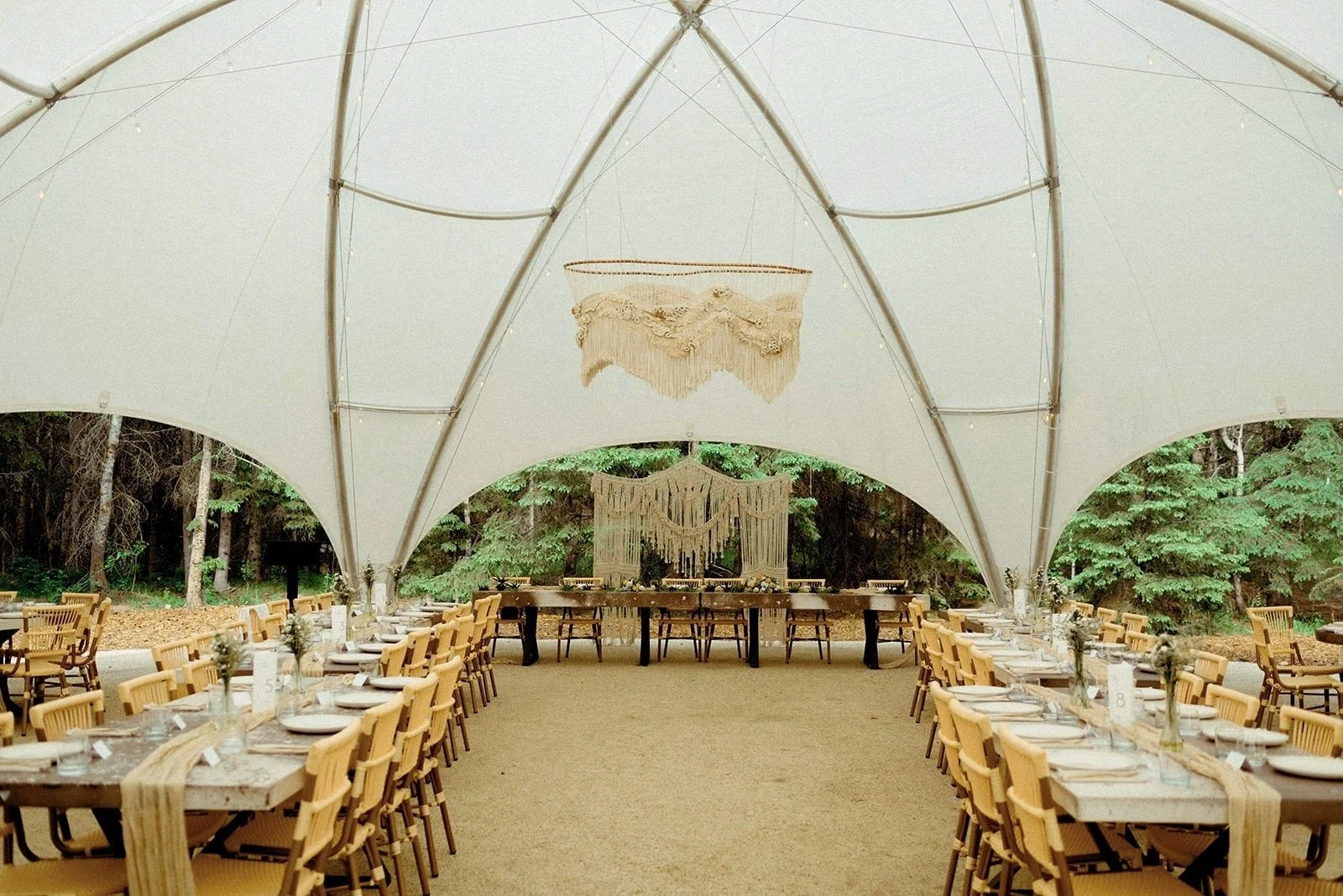 Elegant reception tent with long wooden tables set for dining. A large macrame chandelier hangs above, creating a cozy, bohemian ambiance amidst the surrounding forest.