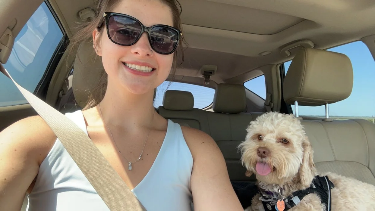 Woman in sunglasses taking a car selfie with a small fluffy dog.