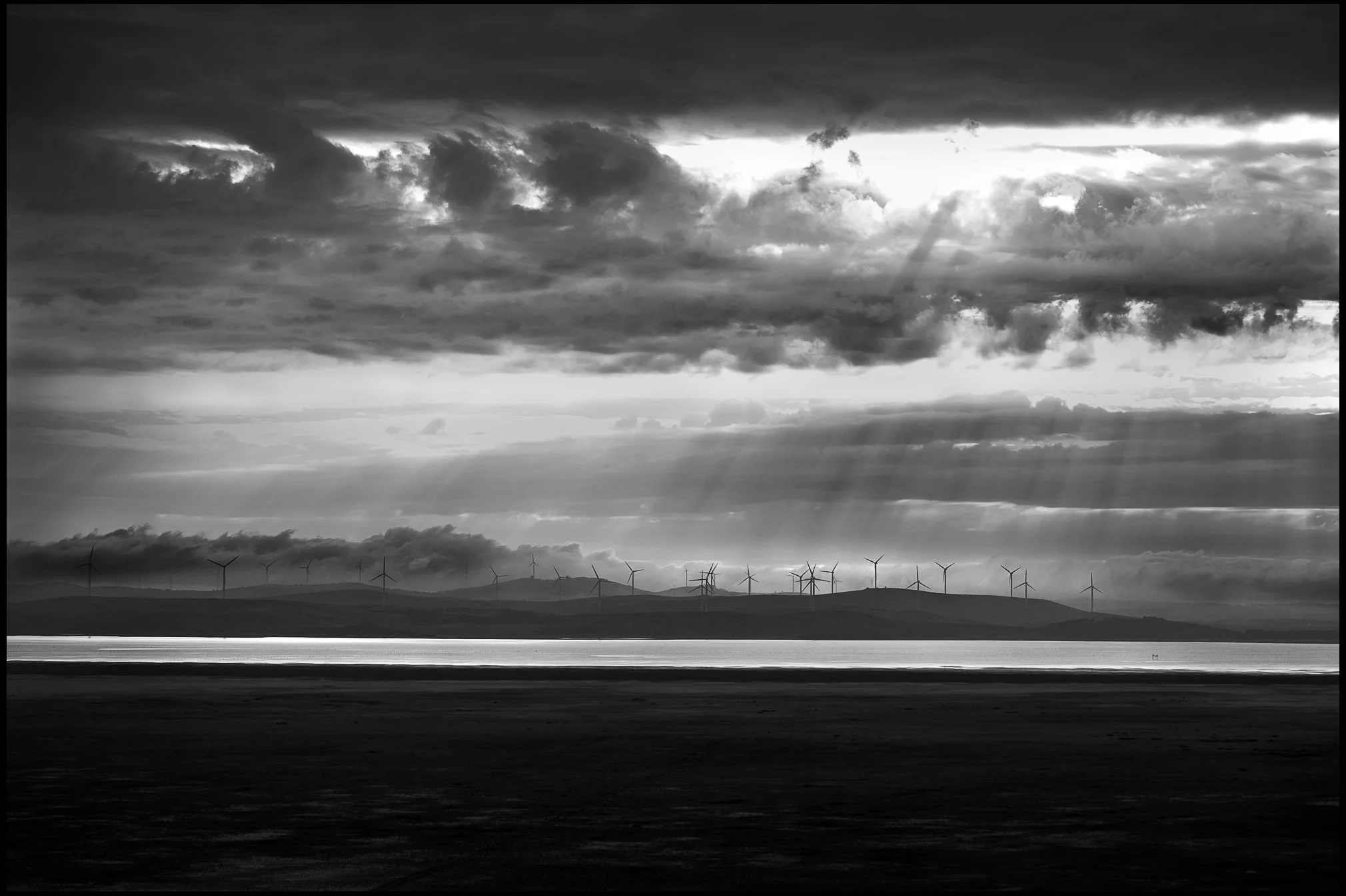 Black and white landscape with a body of water, distant hills, and wind turbines on the horizon under dramatic cloudy sky with sun rays breaking through.