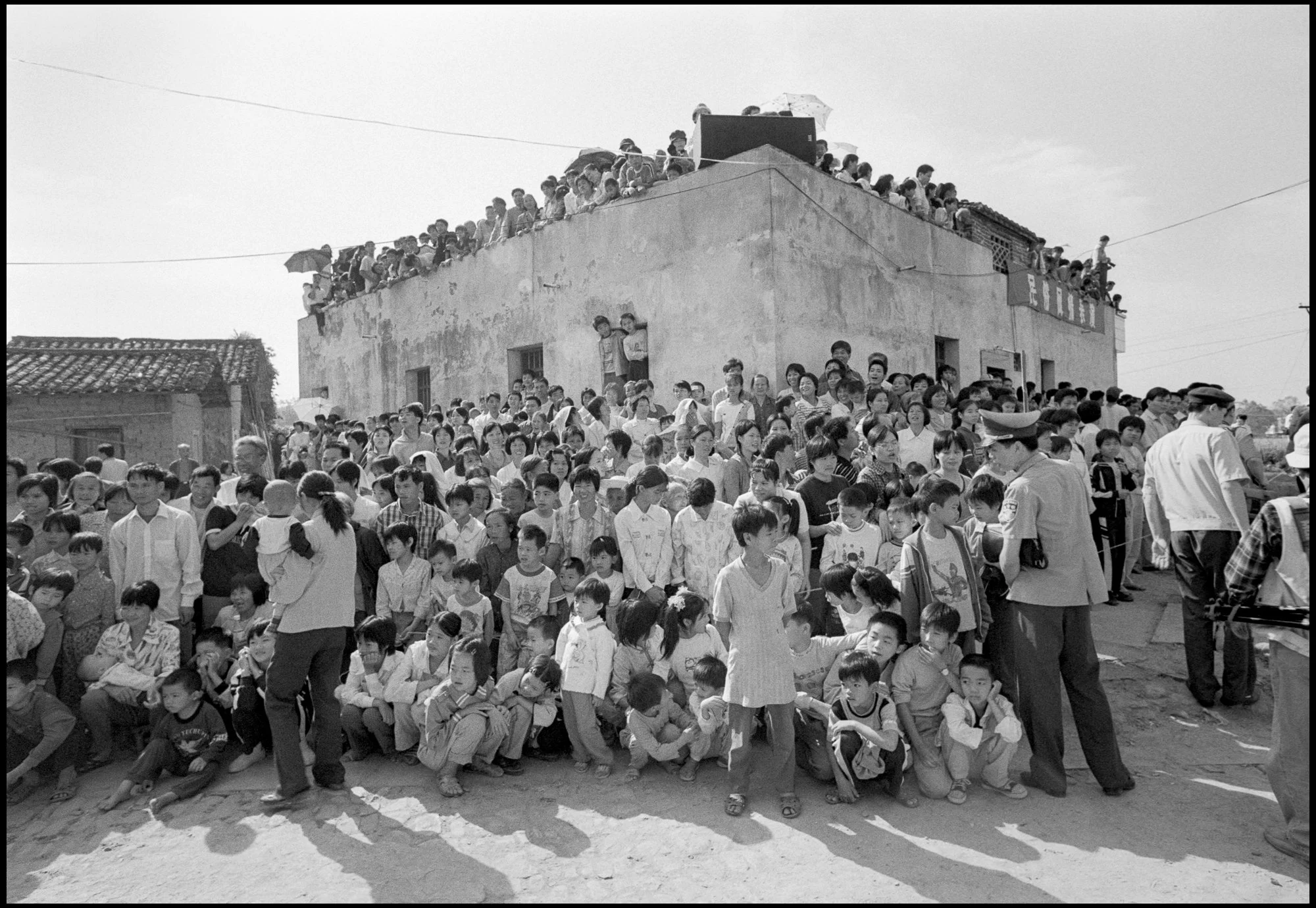 A large crowd of children and adults gathered outside a building, with some people sitting and others standing, in an outdoor setting. There are police officers present, and many people are observing from the rooftop. The photo is in black and white.